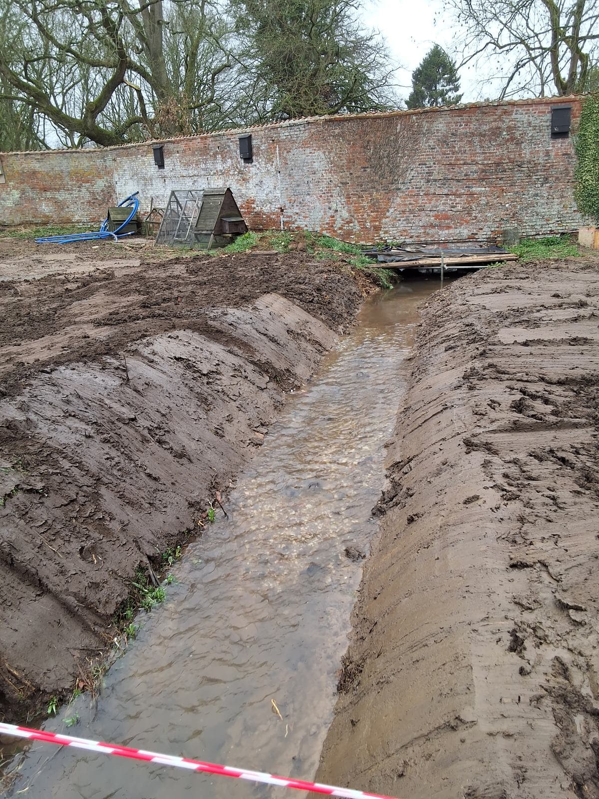 Open trench alongside a brick boundary wall, holding water before pipe laying